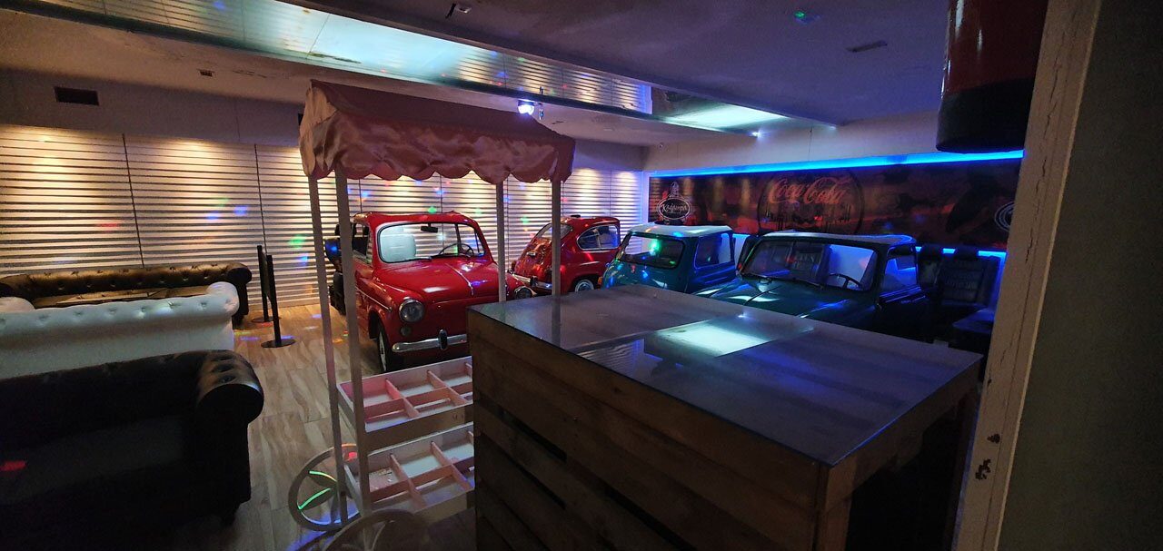 La barra de madera con encimera metálica, y detrás los coches clásicos junto al mural iluminado